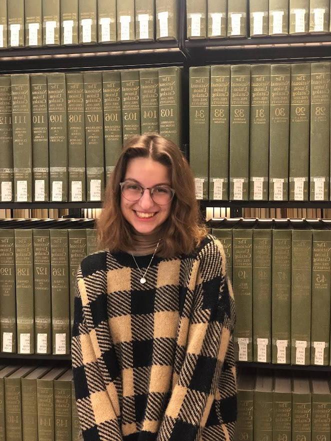 Woman with black and tan checkered shirt and glasses standing in front of book shelf